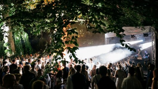 Viele Menschen stehen im Lichtkegel vor der Bühne Baumsaal, über ihnen lange grüne Äste.